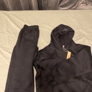 Svart Essentials Fear of God tracksuit - Svart tracksuit från Essentials Fear of God med broderad logga på bröstet och patch på huvan. Klassisk oversized passform, mjukt material och stor huva. Perfekt för en chill och stilren look.