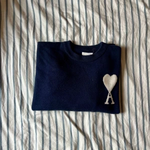 Mörkblå stickad tröja från Ami Paris - Säljer en mörkblå stickad tröja från Ami Paris med deras ikoniska vita hjärta och A på bröstet. Tröjan har rund hals, långa ärmar och breda ribbade muddar vid ärmslut och nederkant. Perfekt för dig som gillar stilrena och trendiga plagg.