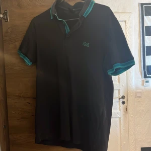 Svart Hugo Boss pikétröja med turkosa detaljer - Snygg svart pikétröja från Hugo Boss med turkosa detaljer på krage och ärmslut. Klassisk polo med korta ärmar och broderad logga på bröstet. Perfekt för en clean och stilren look.