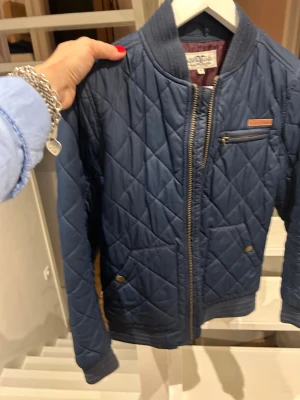 Blå bomberjacka Hampton Republic - Quiltad bomberjacka i mörkblått från Hampton Republic med guldfärgad dragkedja och ribbade muddar. Jackan har en liten bröstficka med dragkedja och två snedställda fickor med tryckknappar. Insidan är fodrad i vinrött och materialet känns lätt och skönt.