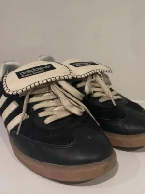 Adidas Wales Bonner svart/vit sneakers - Adidas Wales Bonner sneakers med svart ovandel, vita ränder och detaljer, samt beige snörning. Skorna har en unik, broderad plös med Wales Bonner-logga och en klassisk gummisula i brunt. Materialmix av mocka och skinn ger en retro och cool vibe.
