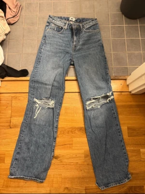 Blå raka jeans med slitna knän - Säljer ett par blå jeans med raka ben och slitna detaljer över båda knäna. Klassisk femficksmodell med normal passform och hög midja. Perfekta för en avslappnad och trendig look.