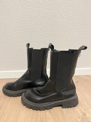 Svarta chunky chelsea boots - Säljer ett par svarta chunky chelsea boots med grov platt sula och dragkedja baktill. Bootsens ovandel är i blankt skinn och har elastiska paneler på sidorna för enkel påtagning. Perfekt för dig som gillar en edgy och stilren look.