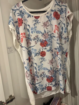 Vit blommig topp från Esmara - Säljer en vit topp från Esmara med färgglatt blommigt mönster i rött, blått och ljusrosa. Toppen har rund halsringning och är ärmlös, perfekt för en fräsch och somrig look. Materialet känns lätt och luftigt, vilket gör den skön att bära under varma dagar.