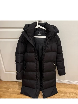 Lång vinterjacka unisex  - Säljer en svart lång pufferjacka . Jackan har en hög krage, huva och dragkedja framtill samt två fickor med dragkedja. Den är vadderad och har en clean, stilren look som håller dig varm under kalla dagar. Storlek Medium säljs för 800kr men priset går att diskutera.