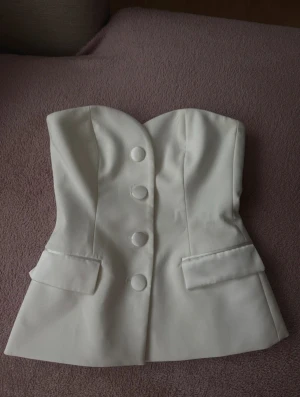 Vit korsett-topp med knappar - Säljer en snygg vit korsett/kostym-topp med axelbandslös design och markerad sweetheart-ringning ifrån Zara, i storlek S. Toppen har stora dekorativa knappar framtill och två fejkade fickdetaljer. Perfekt för utgångar!