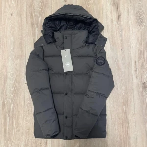 Mörkgrå dunjacka från Canada Goose - Snygg mörkgrå dunjacka från Canada Goose med huva och svart logotyp-patch på ärmen. Jackan har dragkedja och knappar framtill, quiltad design och flera praktiska fickor. Perfekt för kalla dagar och har en clean, modern look.