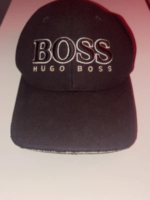Svart Hugo Boss keps med broderad logga - Snygg svart keps från Hugo Boss med stor broderad BOSS-logga framtill och justerbart spänne bak. Kepsen är tillverkad i ett slitstarkt bomullsmaterial och har klassisk form med böjd skärm. Perfekt för att lyfta din streetwear-look.