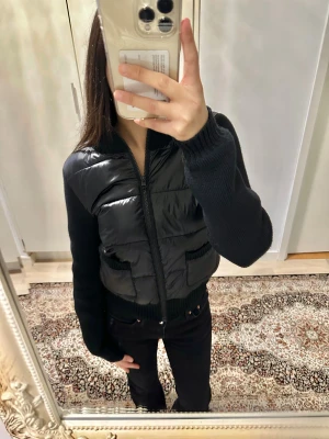 Cardigan med stickade ärmar - Säljer en svart cardigan/jacka med stickade ärmar, rygg och ribbad krage. Jackan har två fickor framtill och dragkedja hela vägen upp. Kroppsdelen är quiltad och glansig medan ärmarna är matta och stickade, vilket ger en snygg kontrast. Storlek M men sitter som XS. Nyskick.