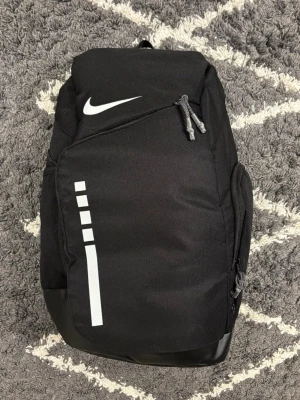 Nike Elite bag - Snygg svart ryggsäck från Nike med vita detaljer och logga. Rymligt huvudfack, flera dragkedjefickor och vadderade axelremmar med coola transparenta partier. Perfekt för skolan, gymmet eller vardagsäventyr. 