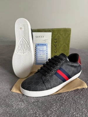 Gucci skor  - Snygga svarta Gucci sneakers med GG-monogram i mörkgrått, vita sulor och klassiska röd-blå ränder på sidan. Baksidan har metallicröd och metallicblå häl i ormskinnsmönster med Gucci-logga. Skorna har snörning och rund tå, tillverkade i skinn och syntet.