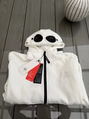 C.P Company vit kofta  - Vit zip-hoodie från C.P. Company. På sidan om dragkedjan finns fickor. Passformen är normal. Märkets logga är tryckt och placerad på bröstet samt att märkets välkända goggles finns på huvan.