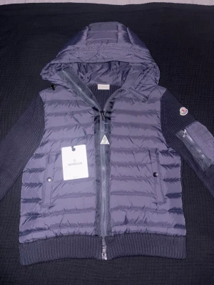 Mörkblå Moncler pufferjacka med huva - Snygg mörkblå pufferjacka från Moncler med stickade ärmar och huva. Jackan har dragkedja framtill, två fickor med knappar och en extra ficka med dragkedja på ärmen. Materialmix av polyester och stickat ger en unik look. Perfekt för kyliga dagar.