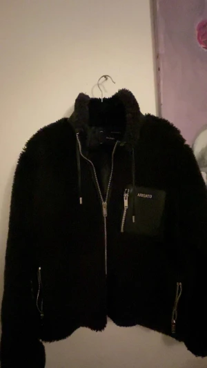 Svart teddyjacka från Axel Arigato - Svart teddyjacka från Axel Arigato med fluffig yta och coola detaljer. Jackan har dragkedja framtill, snörning vid kragen och en snygg ficka med logotyp och dragkedja på bröstet. Perfekt för dig som gillar streetstyle och vill ha något unikt. Priset kan diskuteras 🥂
