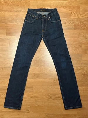 Nudie Jeans Co W28 L32 - Snygga mörkblå jeans från Nudie Jeans Co i klassisk rak modell. Jeansen har kontrastsömmar, fem fickor och läderpatch med logga bak. Tillverkade i slitstarkt bomullsjeans-tyg, perfekta för en clean och stilren look.