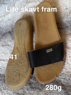 Svarta sandaler stl 41 - Säljer ett par stilrena sandaler i storlek 41. De har en svart, perforerad rem över foten och en ljusbrun sula med blommönster undertill. Perfekta för varma dagar och enkla att matcha med olika outfits.🌸 Lite skav fram. Storlek 41