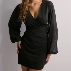 Svart omlottklänning med v-ringning - Supersnygg svart kort omlottklänning med djup v-ringning och långa, transparenta ärmar. Klänningen har en figurnära passform och markerad midja som ger en riktigt chic siluett. Nyår