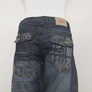 Y2k wide jeans med tryck från Saroe - Unika blå jeans med breda ben och coolt tryck i grått över hela byxan. Modellen har stora fickor med lock och knapp bak, samt detaljerade sömmar på benen. Längd : 101cm Midjemått tvärs över : 36 cm Benvidd: 22 cm 
