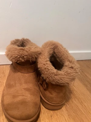 Bruna fluffiga boots med rund tå - Mysiga boots i brun mocka med fluffig päls runt skaftet och rund tå. Snyggt spänne på sidan och platt sula som ger skön komfort. Perfekta för kalla dagar när du vill ha både stil och värme.