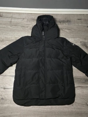 Svart dunjacka från Canada Goose - Svart dunjacka från Canada Goose med klassisk rund logotyp på ärmen. Jackan har dragkedja och är tillverkad i slitstarkt syntetmaterial med dunfyllning. Perfekt för kalla dagar och har praktiska axelremmar på insidan för att bära jackan smidigt.