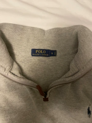 Grå half zip tröja från Polo Ralph Lauren - Säljer en grå långärmad tröja från Polo Ralph Lauren i storlek S. Tröjan har en half zip med läderdetalj, hög krage och det klassiska broderade Polo-logot på bröstet. Materialet är mjukt och stickat, perfekt för kyliga dagar.