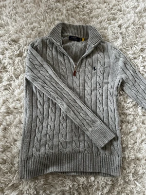Grå stickad half zip-tröja Polo Ralph Lauren - Snygg grå kabelstickad tröja från Polo Ralph Lauren med half zip och ribbad krage. Perfekt för dig som gillar varma snygga tröjor till vintern. Mycket bra skick. Hör av er vid frågor eller om pris. Den är äkta