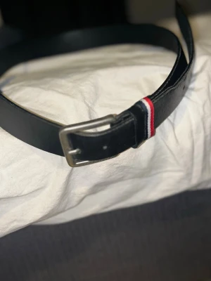 Svart bälte från Jack & Jones - Snyggt svart bälte från Jack & Jones med metallspänne och en cool detalj i form av en röd, vit och blå rand vid spännet. Bältet är tillverkat i skinn och har en stilren, klassisk form som passar till jeans och chinos. Väldigt bra skick, och inte använd så mycket. Köpt för 289 kr. Pris kan diskuteras!