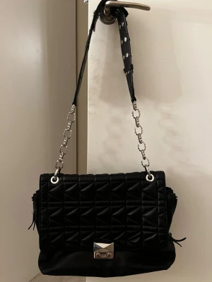 Svart quiltad axelväska Karl Lagerfeld - Stilren svart axelväska från Karl Lagerfeld i quiltat skinn med silverfärgad kedja och coolt spänne framtill. Väskan har detaljer i metall, justerbart axelband och innerfack med dragkedja. Perfekt för dig som vill ha en edgy och trendig look.