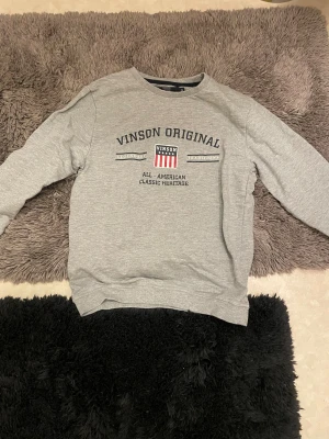 Grå sweatshirt från Vinson Original - Säljer en grå sweatshirt från Vinson Original med tryck framtill i svart, vitt och rött. Tröjan har rund halsringning och långa ärmar. Skön och enkel stil med klassisk amerikansk vibe, perfekt till jeans eller joggers. Priset kan sänkas vid diskussion 