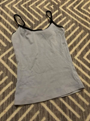 Ljusblå linnetopp med svarta kanter - Säljer en stilren ljusblå linnetopp med tunna axelband och svarta kanter runt halsringningen. Toppen har en enkel, rak passform.