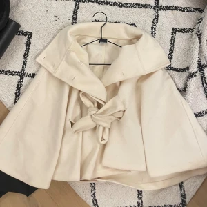 Beige kappa med bälte från Gina Tricot - Snygg beige kappa från Gina Tricot i storlek XS/S. Jackan har bred krage, knappar framtill och ett knytbälte i midjan. Materialet är mjukt och känns lyxigt, perfekt för dig som vill ha en stilren look med lite extra edge.