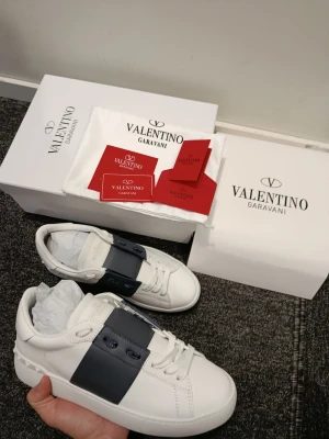 Valentino Garavani vita sneakers - Snygga vita sneakers från Valentino Garavani med bred mörkblå rem över snörningen och rund tå. Skorna har en platt sula med mönstrad undersida och är tillverkade i skinn. Ikonisk logga på plösen och sula. Perfekt för dig som gillar stilrena designer.