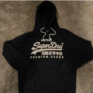 Svart Superdry hoodie med tryck - Svart hoodie från Superdry med stort vitt tryck på bröstet och japanska tecken. Klassisk känguruficka, vita snören i huvan och mjukt material. Perfekt för en chill och avslappnad stil.