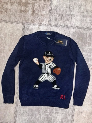 Mörkblå stickad tröja Polo Ralph Lauren - Snygg mörkblå stickad tröja från Polo Ralph Lauren med en stor broderad nalle i basebollkläder på framsidan. Rund hals, långa ärmar och RL-broderi i rött nedtill. Perfekt för dig som gillar sportiga och unika plagg med cool design.