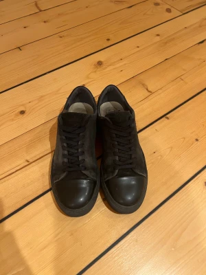 Sneaky Steve svarta  - Snygga svarta sneakers med ovandel i mocka och detaljer i blankt skinn på tå och häl. Klassisk rund tå och platt sula, med svarta snören för en clean look. Perfekt för dig som gillar stilrena och minimalistiska skor.
