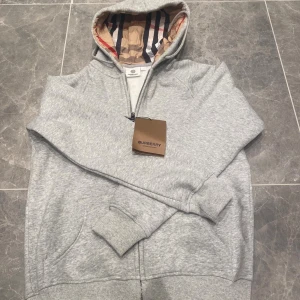 Grå hoodie från Burberry med half zip - Snygg grå hoodie från Burberry med klassiskt rutigt foder i huvan och half zip framtill. Tröjan har långa ärmar, ribbade muddar och fickor på sidorna. Perfekt för dig som gillar stilren design med en lyxig touch. Kan gå ned vid snabb affär.