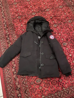 Svart dunjacka Canada Goose XS - Svart dunjacka från Canada Goose i storlek XS. Jackan har huva, flera fickor med lock, dragkedja och stora knappar framtill. Klassisk patch på ärmen och varm vaddering som håller dig cozy hela vintern. Perfekt för dig som gillar streetstyle och vill ha något riktigt fett.