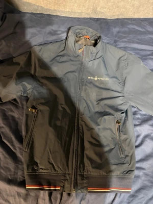 Sail Racing jacka - Snygg mörkblå vindjacka från Sail Racing med hög krage och dragkedja framtill. Jackan har två sidofickor med dragkedja och ribbade muddar med röd och gul rand. Perfekt för dig som gillar sportig stil och vill ha något lätt och funktionellt. Köpt för 2700kr, säljes för 1100kr. Ej använd mycket