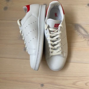 Adidas Stan Smith vita sneakers - Fräscha vita Adidas Stan Smith sneakers med röda detaljer på hälen och tungan. Klassisk låg modell med snörning och perforerade sidor. Skorna har rund tå och platt sula, tillverkade i skinn för en clean och tidlös look.