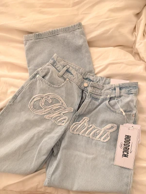 Hoodrich ljusblå wide leg jeans - Säljer ett par ljusblå jeans från Hoodrich med breda ben och broderad logga framtill. Jeansen har normal passform, klassisk femficksdesign och en snygg slitning på ena benet. Materialet är jeans och midjan är midwaist. Perfekt för dig som gillar streetstyle.
