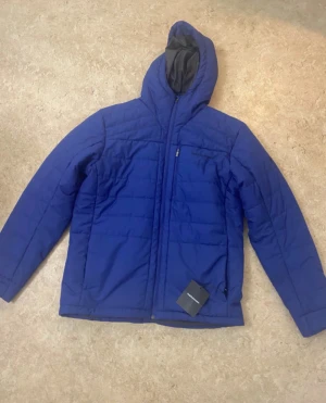 Blå pufferjacka från Peak Performance - Säljer en blå pufferjacka från Peak Performance med huva och hel dragkedja framtill. Jackan har quiltad design och är tillverkad i syntetmaterial, perfekt för kalla dagar. Snygg och enkel stil med diskret logga på bröstet.