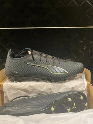 Puma Ultra svarta fotbollsskor - Säljer ett par svarta Puma Ultra fotbollsskor med snygga neongula detaljer och logga. Skorna har snörning och en lätt, syntetisk ovandel med textur för bättre bollkontroll. Sulan har fasta dobbar för gräsplan och en sportig, strömlinjeformad design.