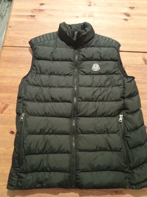 Svart dunväst från Moncler - Svart dunväst från Moncler med quiltad design och hög krage. Västen har två fickor med dragkedja framtill och ett broderat Moncler-märke på bröstet. Insidan har en cool serietidningsetikett och en innerficka med Moncler-logga. Perfekt för lager-på-lager under kyliga dagar.