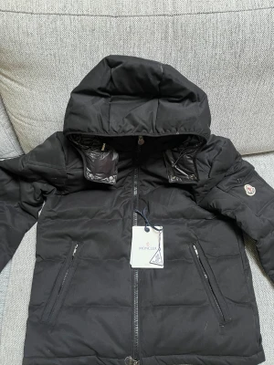 Svart Moncler dunjacka med huva - Säljer min lillebrors jacka. Svart dunjacka från Moncler med stor huva, två snedställda fickor med dragkedja och ikonisk Moncler-logga på ärmen. Jackan har quiltad design och innerfoder med serietidningsmönster. Perfekt för kalla dagar och har en clean, modern look.