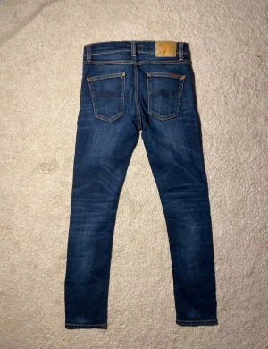 Nudie Jeans Lean Dean mörkblå 27/30 - Snygga Lean Dean jeans från Nudie Jeans i mörkblå tvätt med klassiska orange sömmar. Modellen har smal passform och raka ben, fem fickor och subtila slitningar. Materialet är 99% bomull och 1% elastan för lite stretch. Perfekt för dig som gillar stilrena jeans.