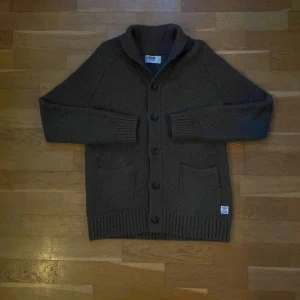 Grön stickad kofta från Dobber - Säljer en snygg mörkgrön stickad kofta/cardigan från Dobber. Den har en klassisk design med knappar framtill och två praktiska fickor. Perfekt för kyligare dagar och ger en stilren look.