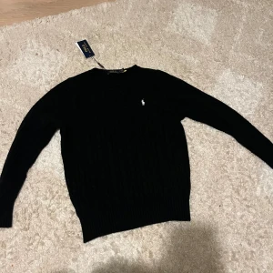 Svart kabelstickad tröja Polo Ralph Lauren - Svart kabelstickad tröja från Polo Ralph Lauren i storlek M. Klassisk design med rund hals och broderad vit logga på bröstet. Tröjan har långa ärmar och ribbade muddar vid ärmslut och nederkant. Perfekt för dig som gillar stilrena och tidlösa plagg. Den är helt ny, aldrig använt denna, prislappen är också på.