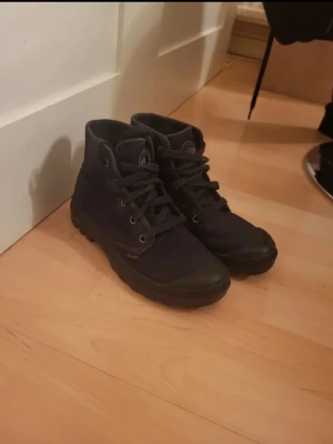 Svarta Palladium boots i canvas - Säljer ett par svarta boots från Palladium i slitstark canvas med grov sula och snörning. Skorna har rund tå och förstärkt tåparti i gummi. Klassisk logga på sidan och tungt street-vibe. Perfekta för dig som gillar robusta och stilrena skor.