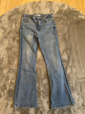 Blå bootcut jeans från Zara, strl 42 - Säljer ett par klassiska blå jeans från Zara i storlek 42. Modellen har hög midja och bootcut-ben som ger en snygg siluett. Jeansen har fem fickor, knapp och dragkedja framtill samt är tillverkade i jeansmaterial med lätt tvättad look.