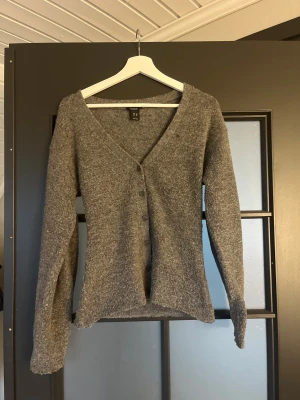 Grå kofta från Lindex med v-ringning - Mysig grå kofta från Lindex i storlek XS. Koftan har v-ringning, långa ärmar och knäppning framtill. Tillverkad i ett stickat material som känns både mjukt och varmt, perfekt för kyligare dagar.